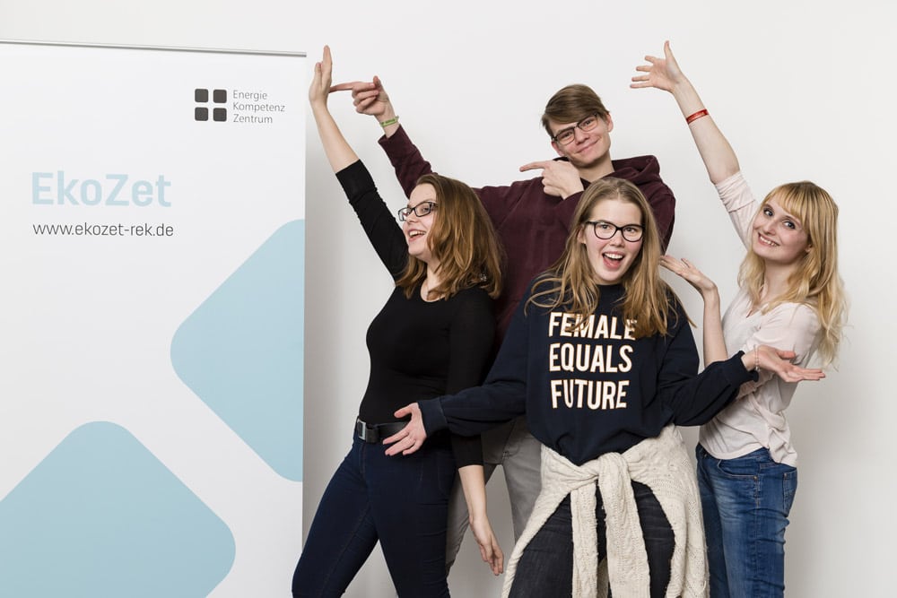Nebem einem Aufsteller mit dem Aufdruck EkoZet - Energie-Kompetenz-Zentrum stehen vier junge Menschen, drei Frauen, ein Mann und zeigen vergnügt auf den Banner. Eine junge Frau schaut direkt in die Kamera, sie trägt ein T-Shirt mit dem Aufdruck " Female equals Future".